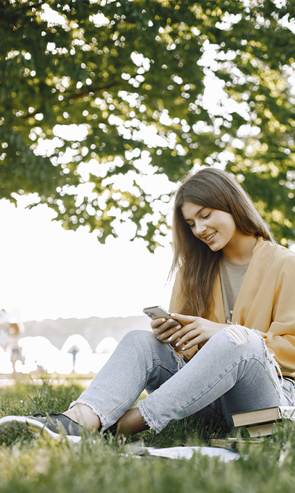 Young woman sitting on the grass surrounded by nature reading our blogs about mental health and connecting with herself.