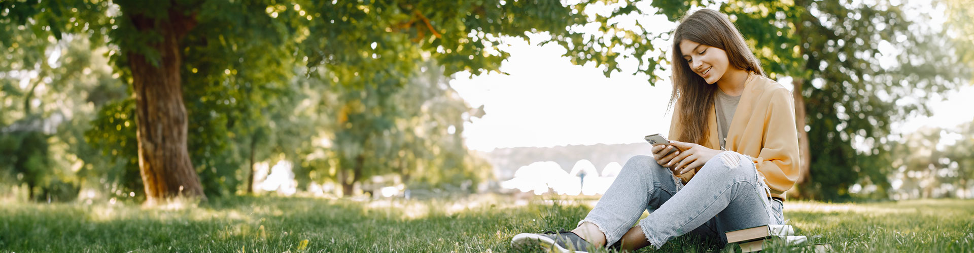 Young woman sitting on the grass surrounded by nature reading our blogs about mental health and connecting with herself.